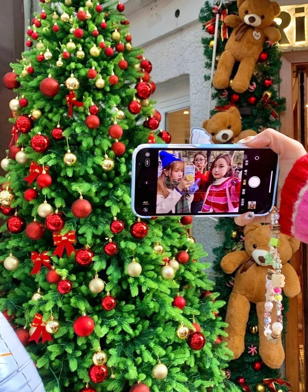 站在聖誕樹前面，其中一部手機開始前置鏡頭，確定所有人在畫面內示意自拍；另一部手機則開啟後置鏡頭，把前面的聖誕樹、自拍的手機畫面同時拍下來。